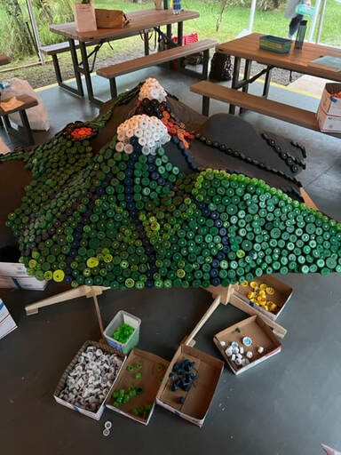 The relief map in process, with one side covered in primarily green caps, with white caps at the summits of Mauna Kea and Mauna Loa. On the floor are boxes of sorted caps with nails through them.